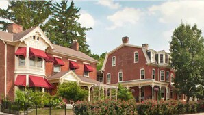 A row of red brick townhouses with white trim and red awnings, surrounded by lush greenery and trees.