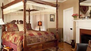 A four-poster canopy bed with a patterned bedspread, a bedside table, a lamp, and a fireplace in the room.