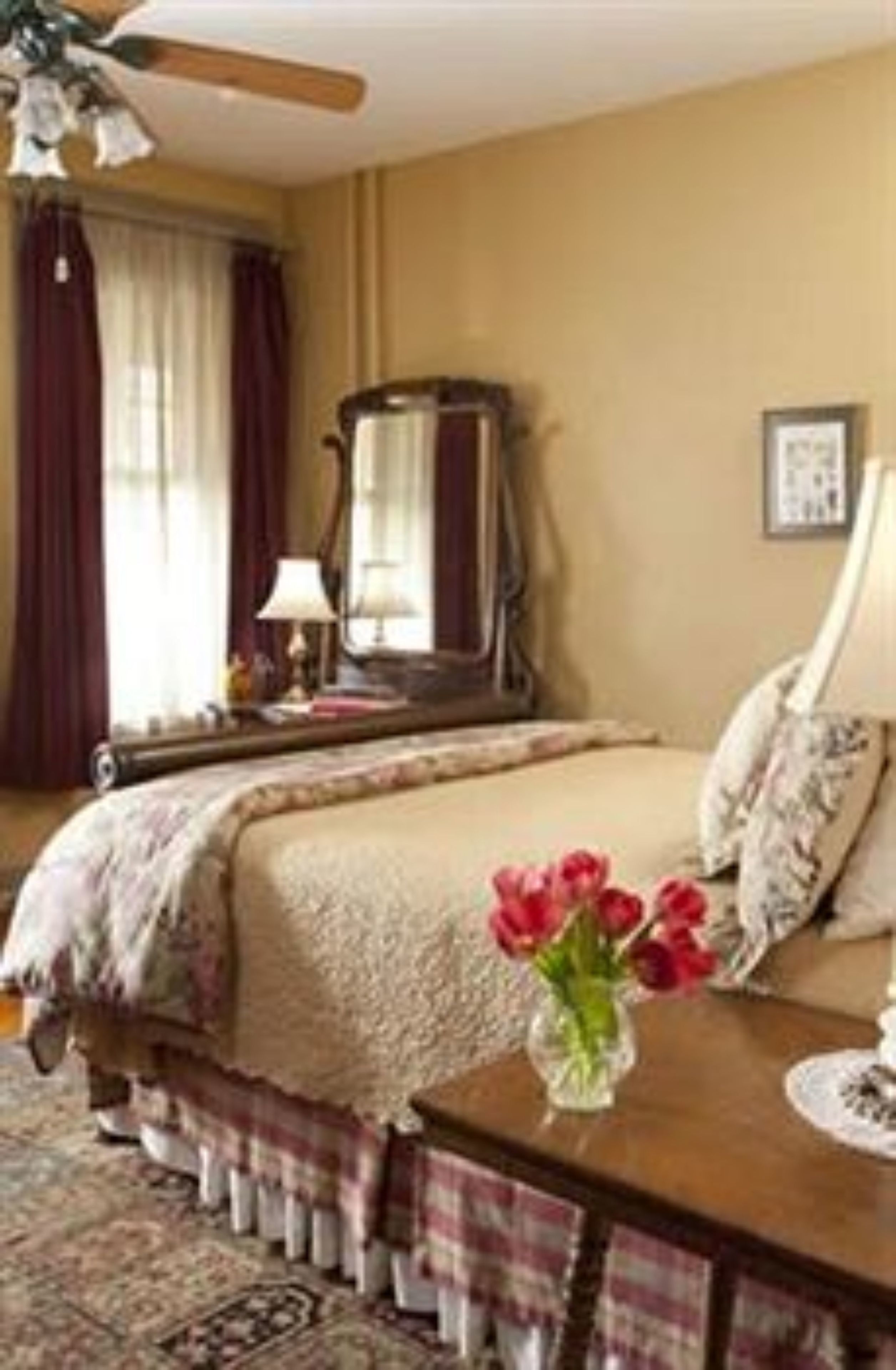 A bedroom with a bed, a nightstand, a lamp, a vase with flowers, and a ceiling fan.