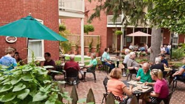 An outdoor patio area with brick flooring, tables, and chairs, surrounded by greenery and a brick building.
