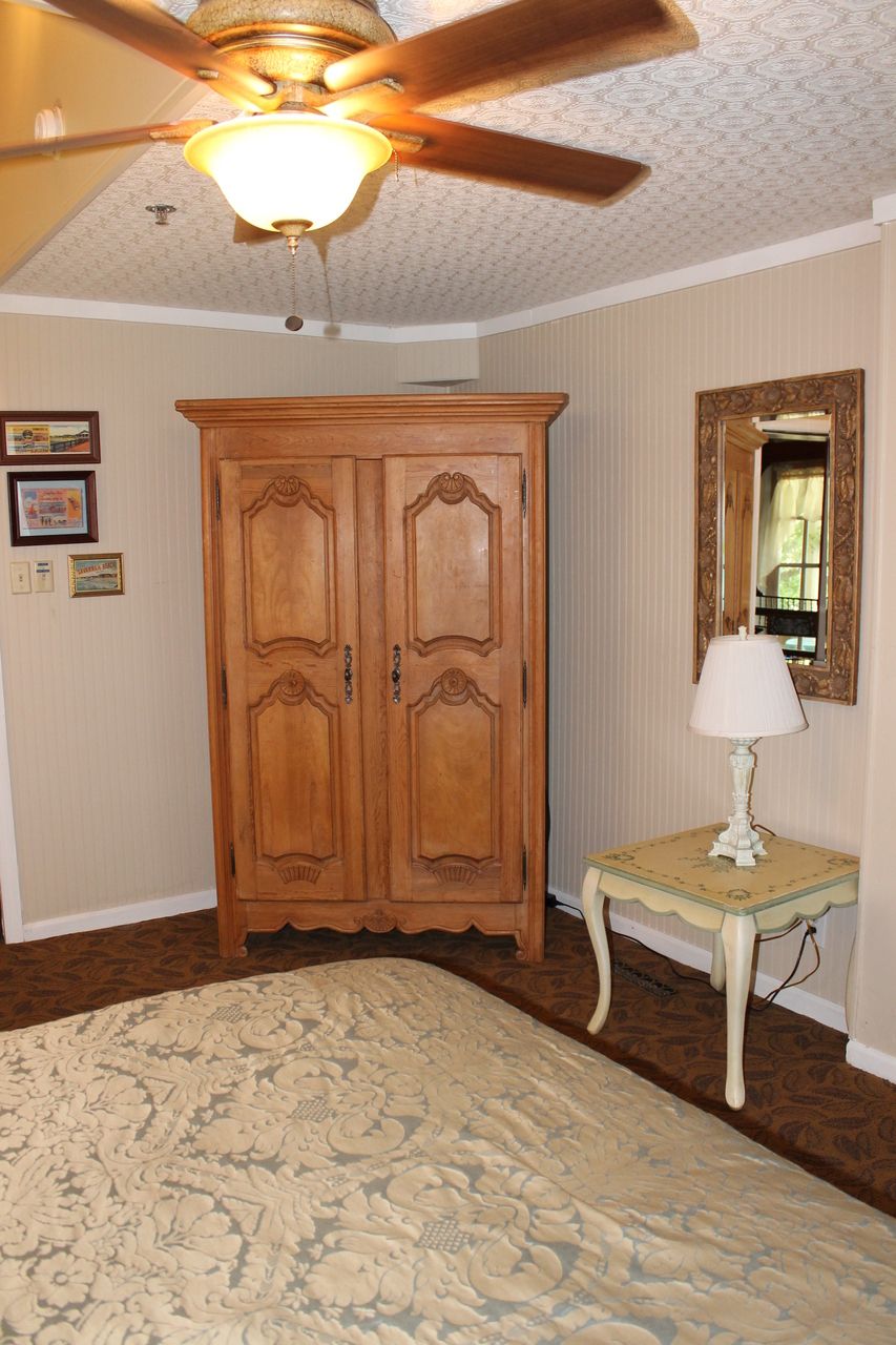Una habitación con un armario de madera, una mesita con una lámpara y un ventilador de techo.