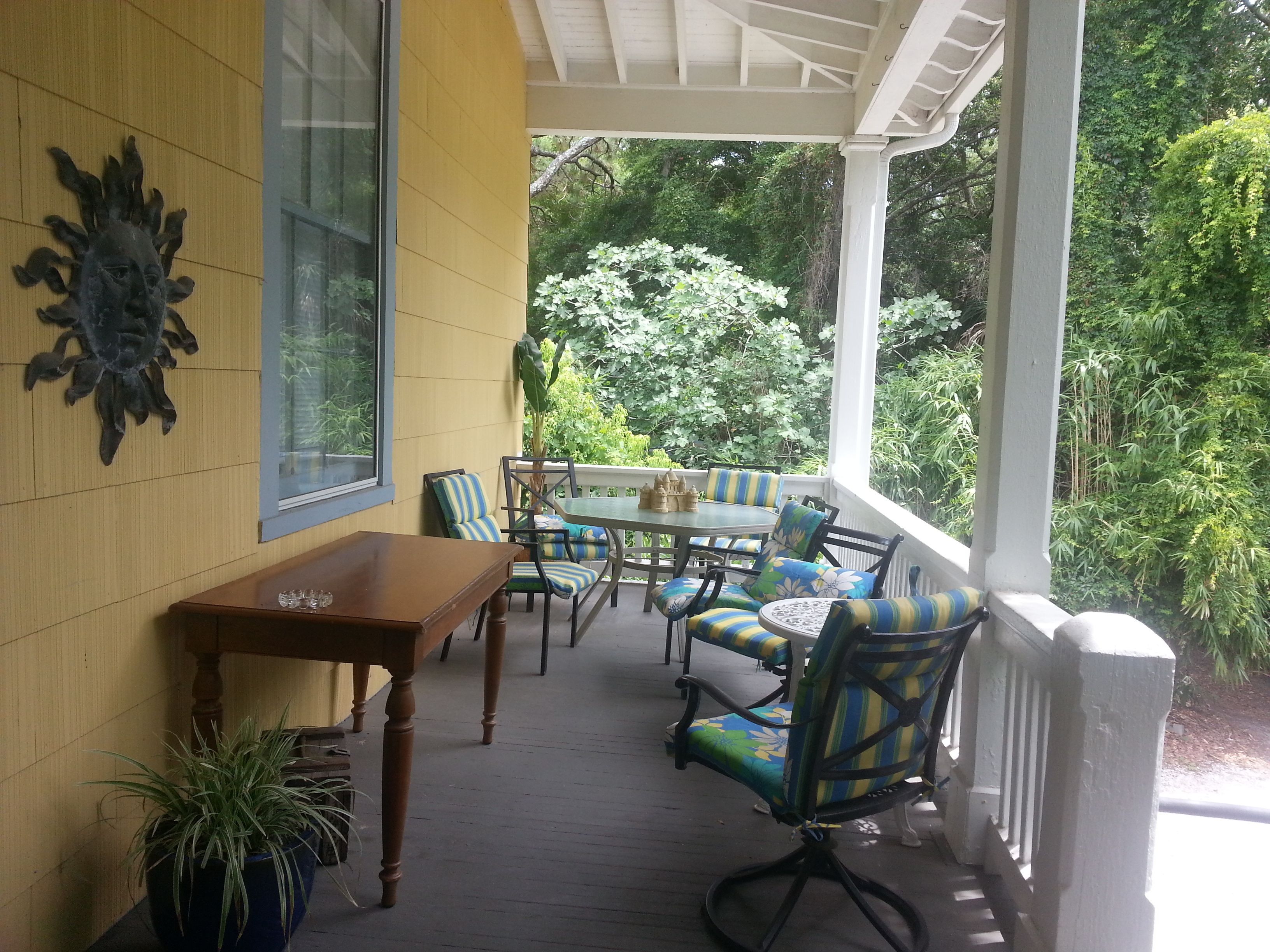 A covered porch with a wooden table and chairs, a potted plant, and a decorative wall piece.