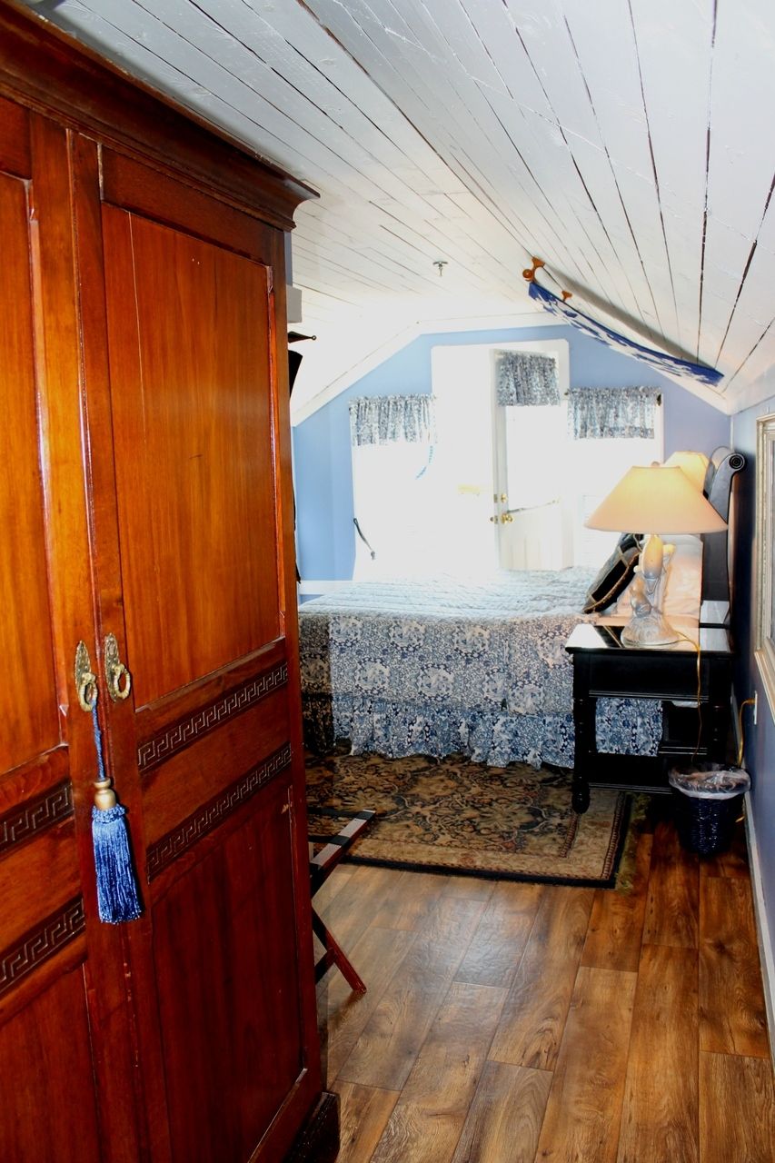 A bedroom with a wooden wardrobe, a bed with a patterned bedspread, a bedside table with a lamp, and a window with curtains.