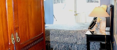 A bedroom with a wooden wardrobe, a bed with a patterned bedspread, a bedside table with a lamp, and a window with curtains.