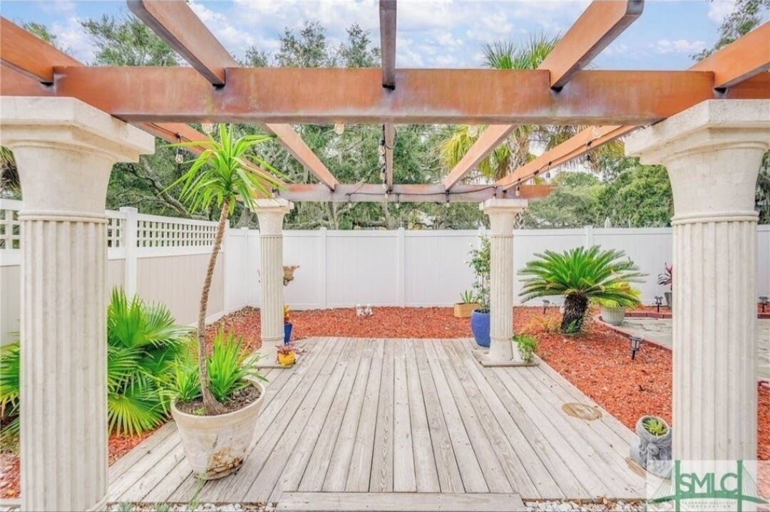 A wooden deck with a pergola structure, surrounded by potted plants and a white fence.