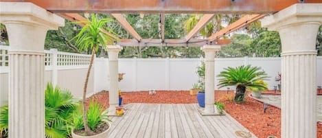 A wooden deck with a pergola structure, surrounded by potted plants and a white fence.