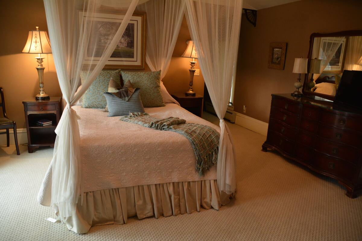 A four-poster bed with a canopy, bedside lamps, a dresser, and a framed picture on the wall.