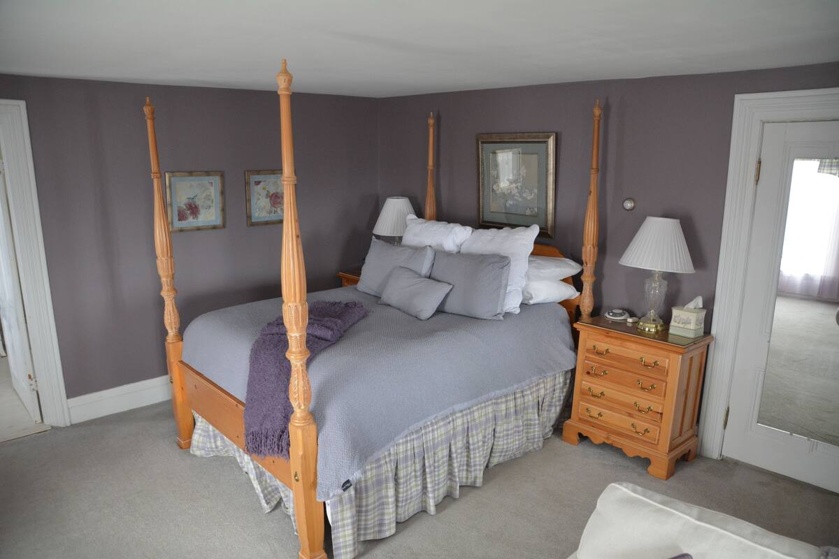 A four-poster bed with a grey bedspread and matching pillows, a wooden nightstand with a lamp, and a small dresser with drawers.