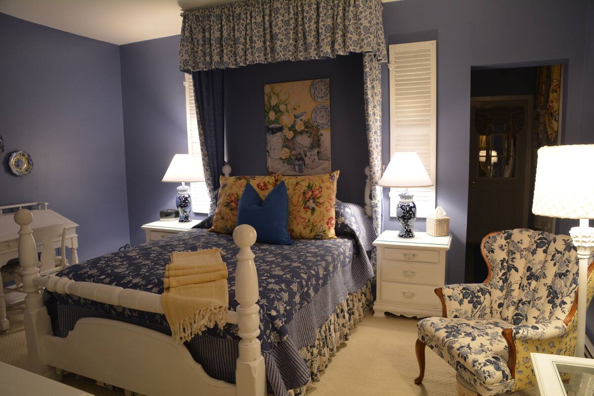 A bedroom with a four-poster bed, floral-patterned bedding, two bedside tables with lamps, a chair with floral upholstery, and a door with a curtain.