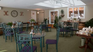 Une pièce spacieuse avec un plafond haut et une lucarne, comprenant un coin repas avec des chaises en métal vert et une table en verre, le tout agrémenté de plantes en pot et d’un tonneau en bois.