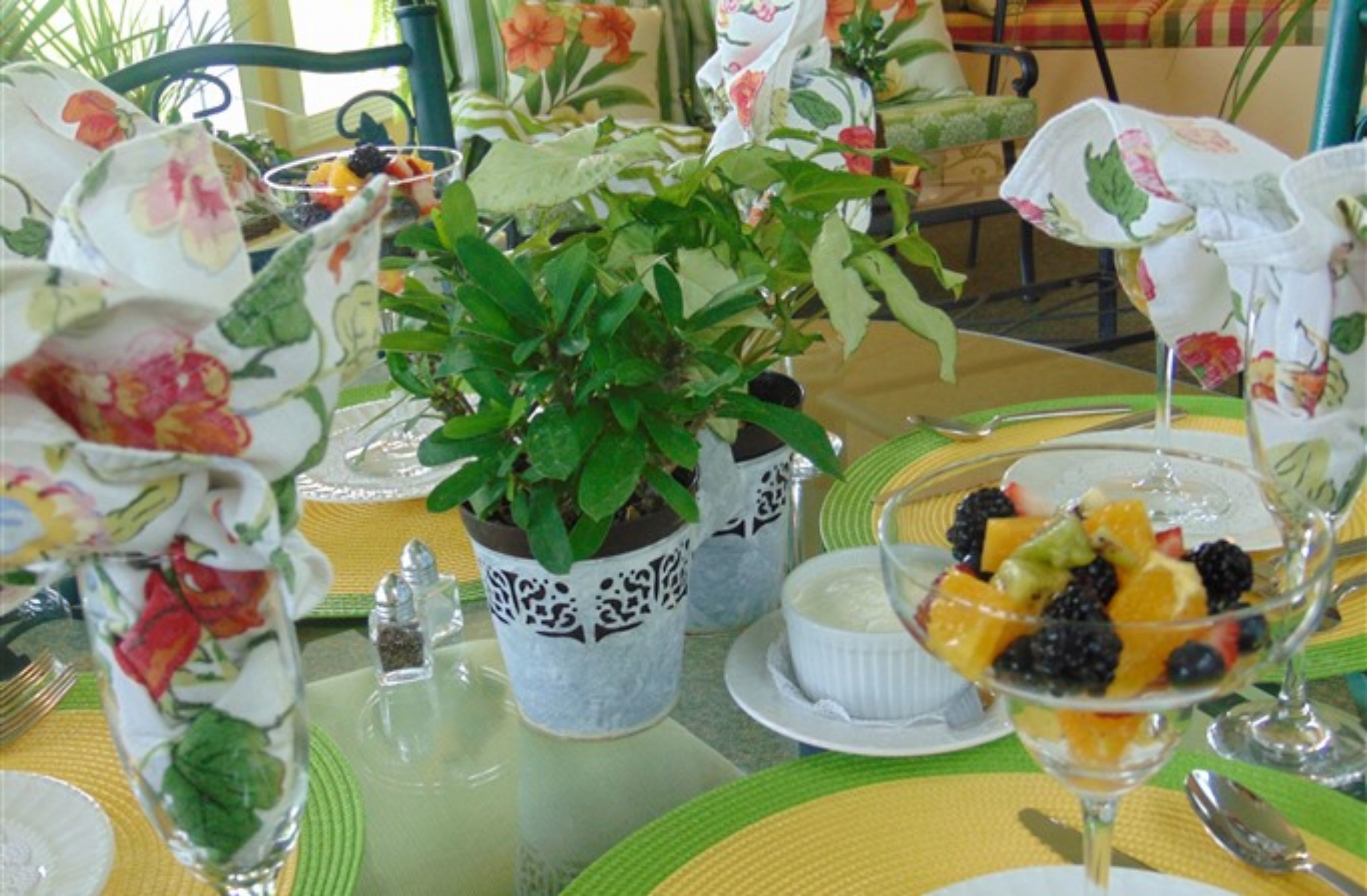 A table set with floral-patterned napkins, a potted plant, a fruit dessert, and a salt and pepper shaker.