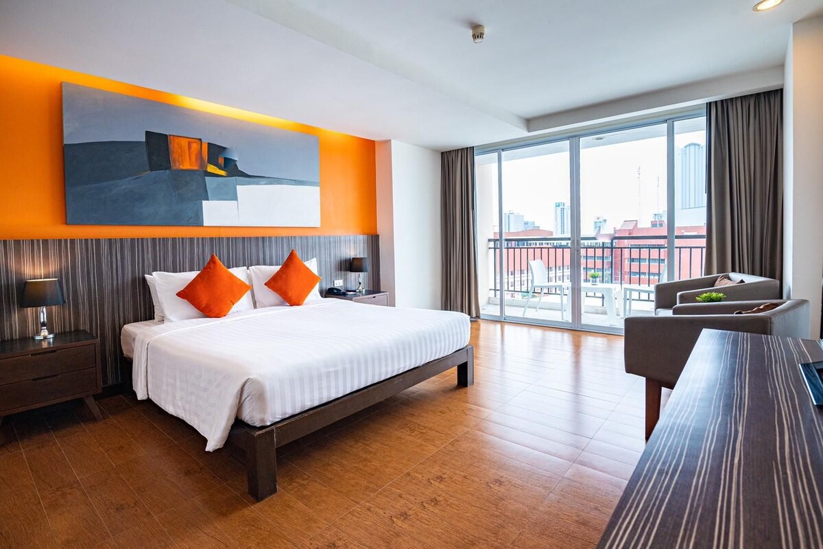 A hotel room with a large bed, a desk, a chair, a nightstand, and a view of the city through large windows.