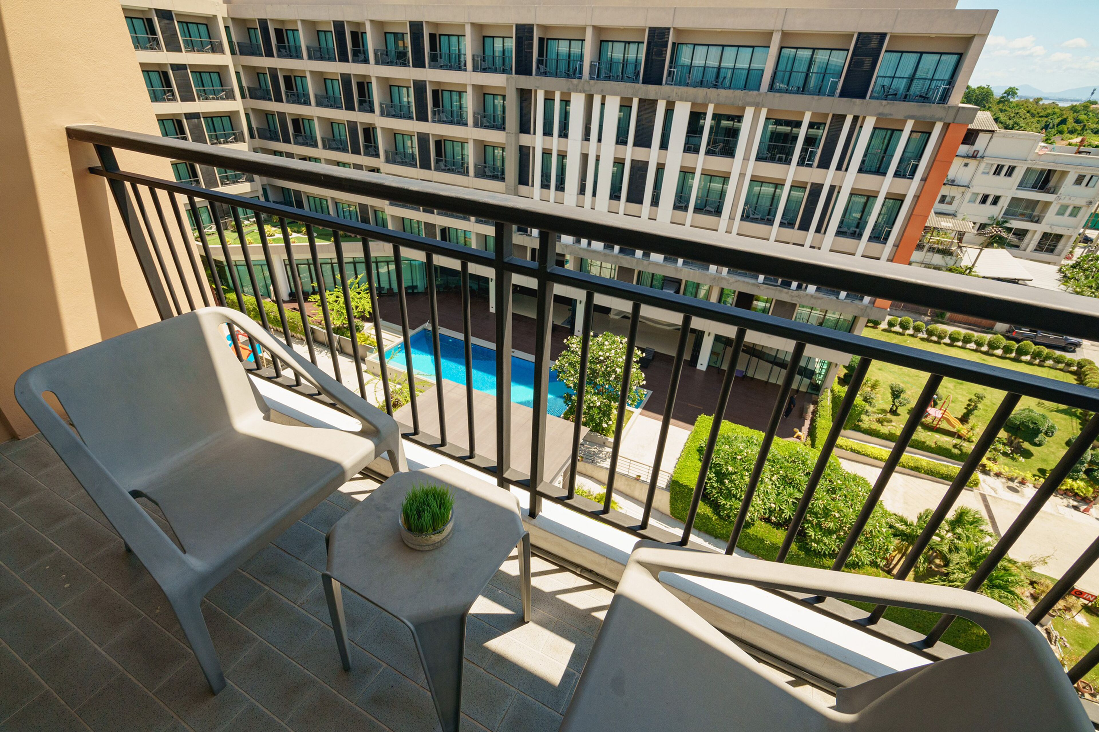 Ein Balkon mit zwei Stühlen, einem kleinen Tisch und einem Topfpflanzen. Im Hintergrund ist ein Schwimmbecken und ein mehrstöckiges Gebäude mit Balkonen zu sehen.