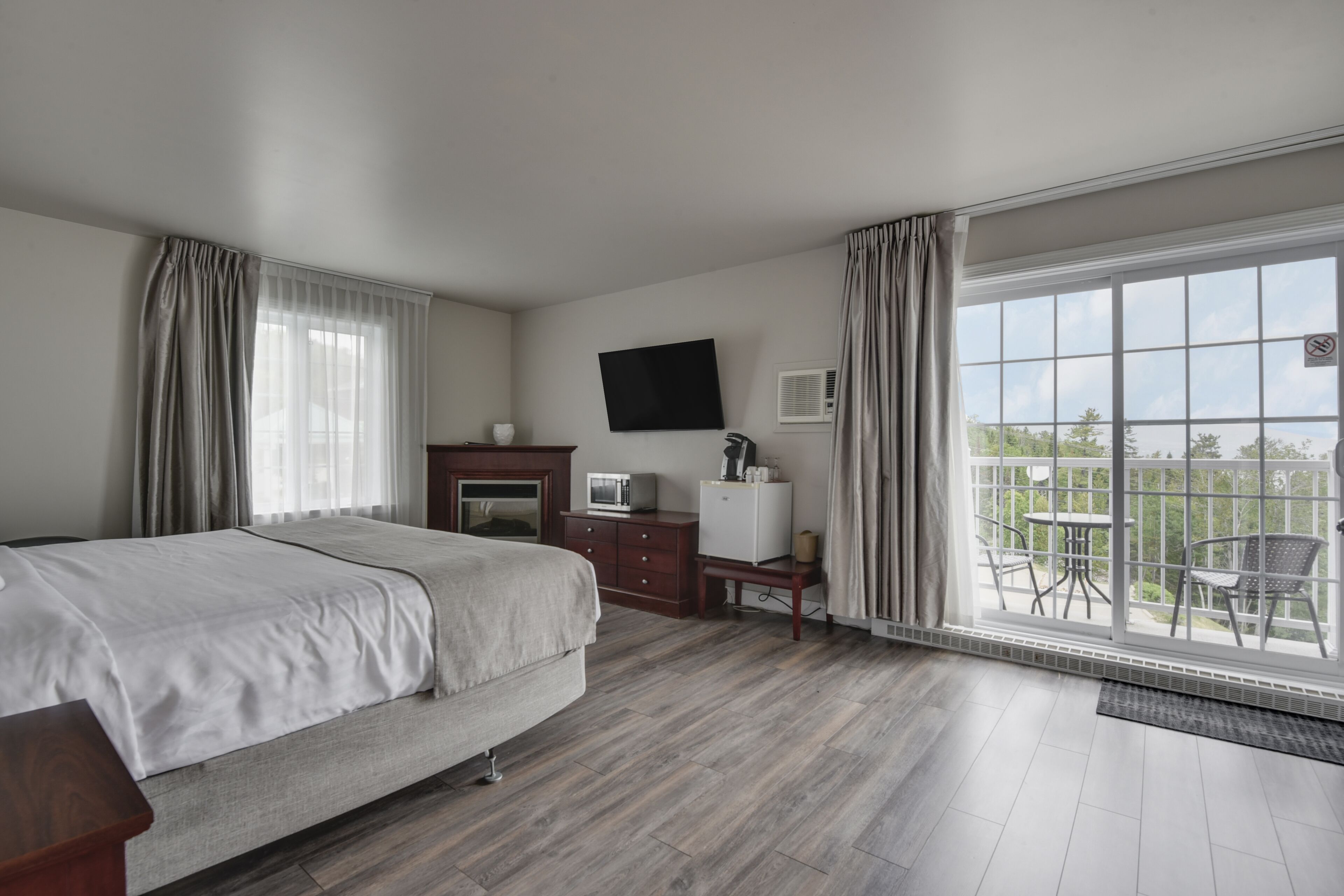 A hotel room with a bed, a flat-screen TV, a small desk with a computer, a window with a view of the outdoors, and a balcony with chairs.