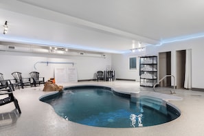 A modern indoor pool area with a curved design, surrounded by black chairs, a small rock feature, and a shelf with towels.
