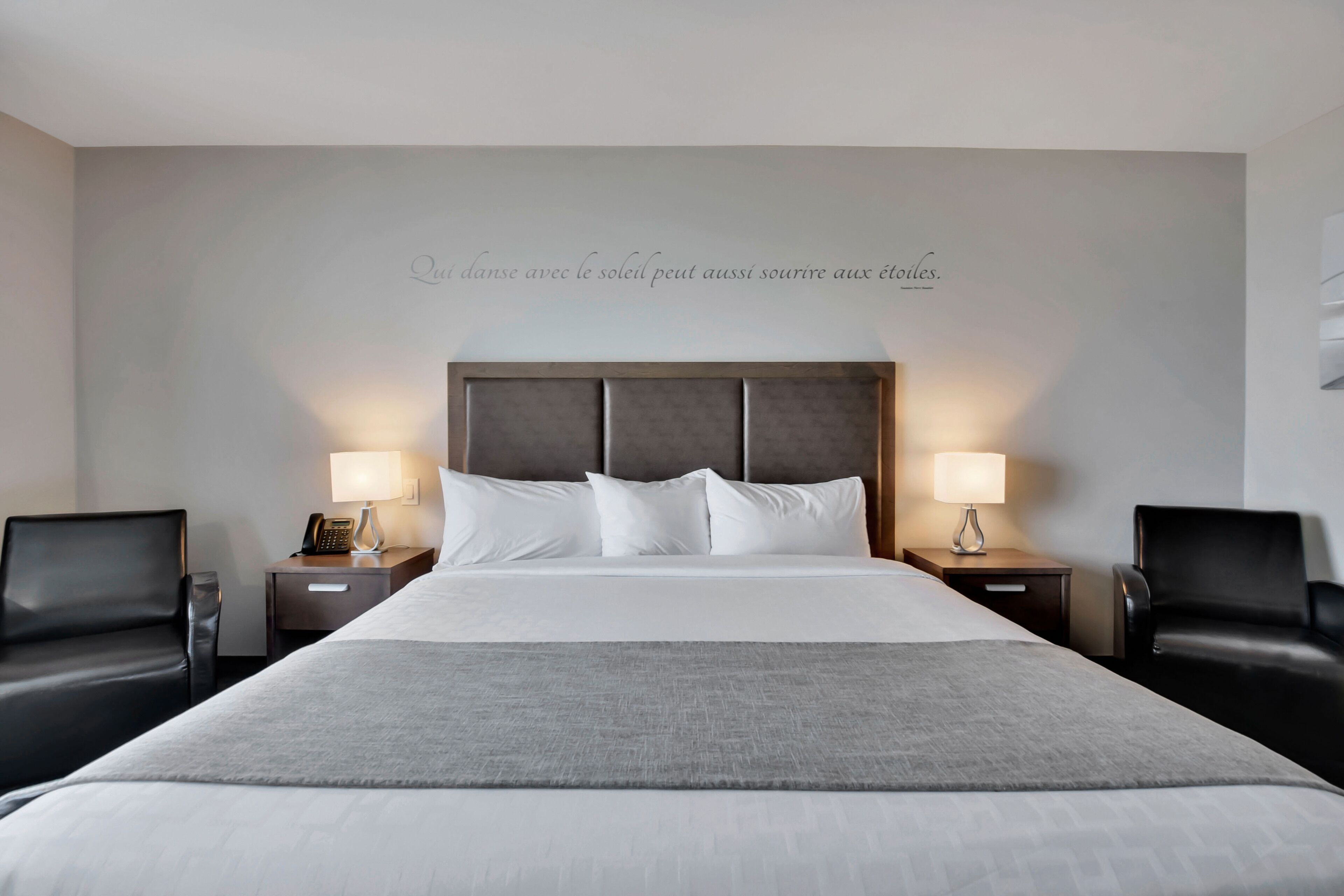 A hotel room with a large bed, two bedside tables with lamps, and two black leather chairs.