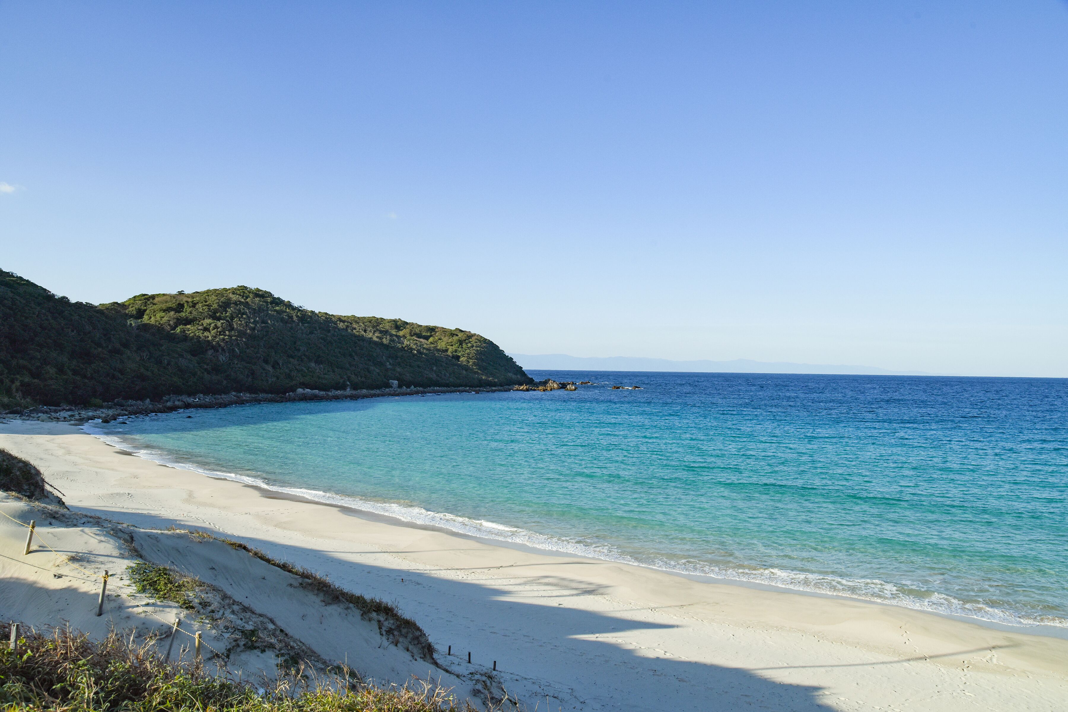 Ein Strand mit klarem türkisblauem Wasser, sandigem Ufer und einer bewaldeten Hügelkette im Hintergrund.