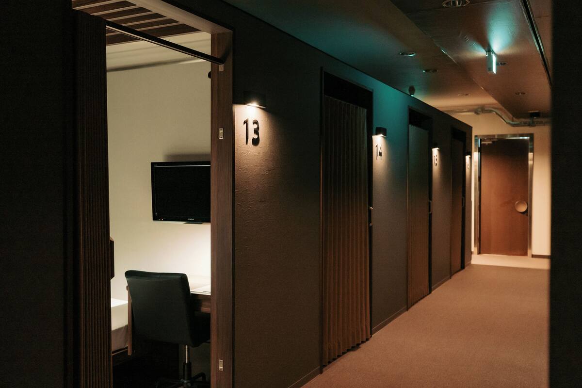 Un couloir d’hôtel avec des portes numérotées, un téléviseur fixé au mur et une chaise dans la chambre.