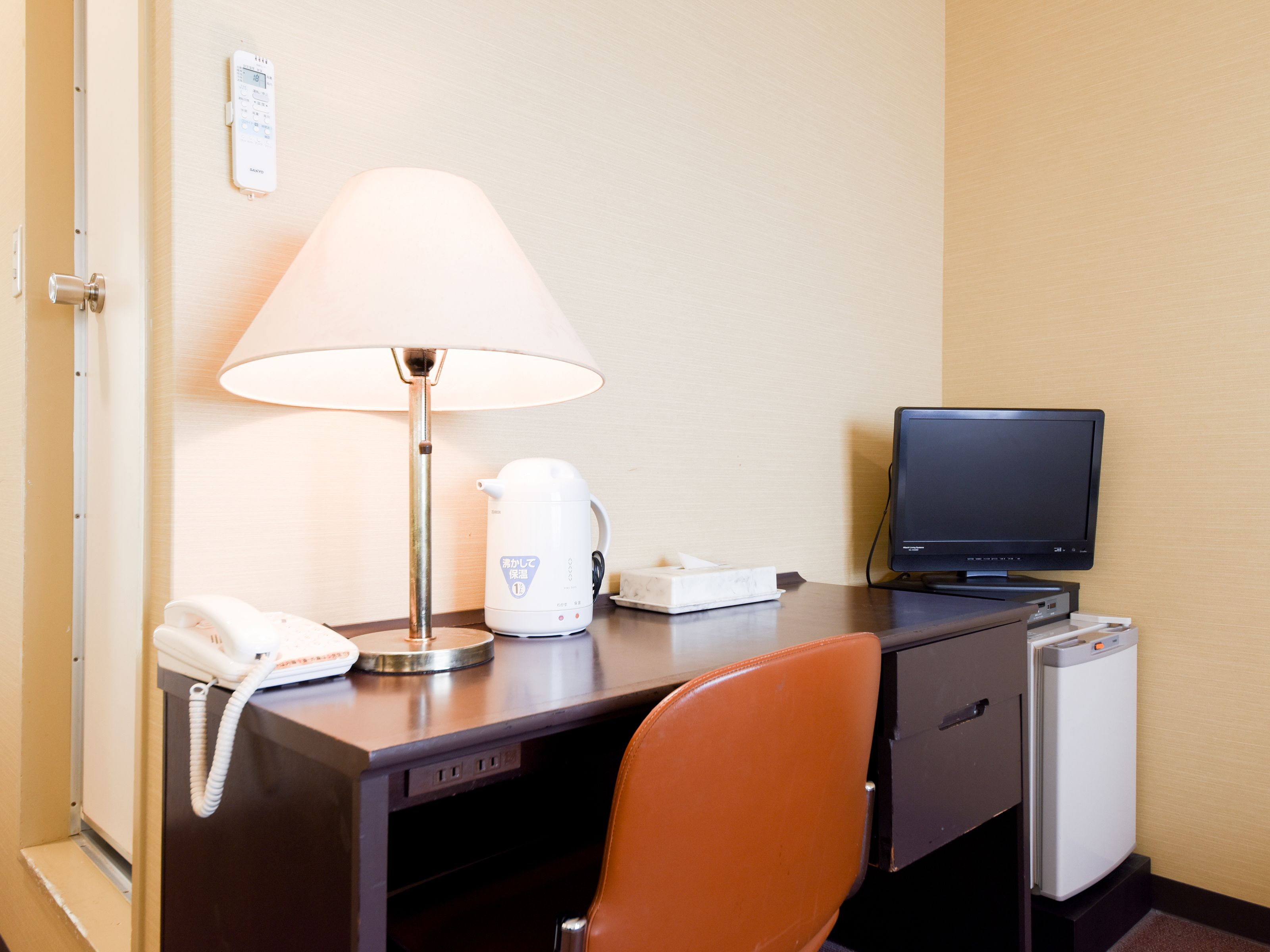 A hotel room with a desk, a lamp, a kettle, a phone, a television, and a refrigerator.
