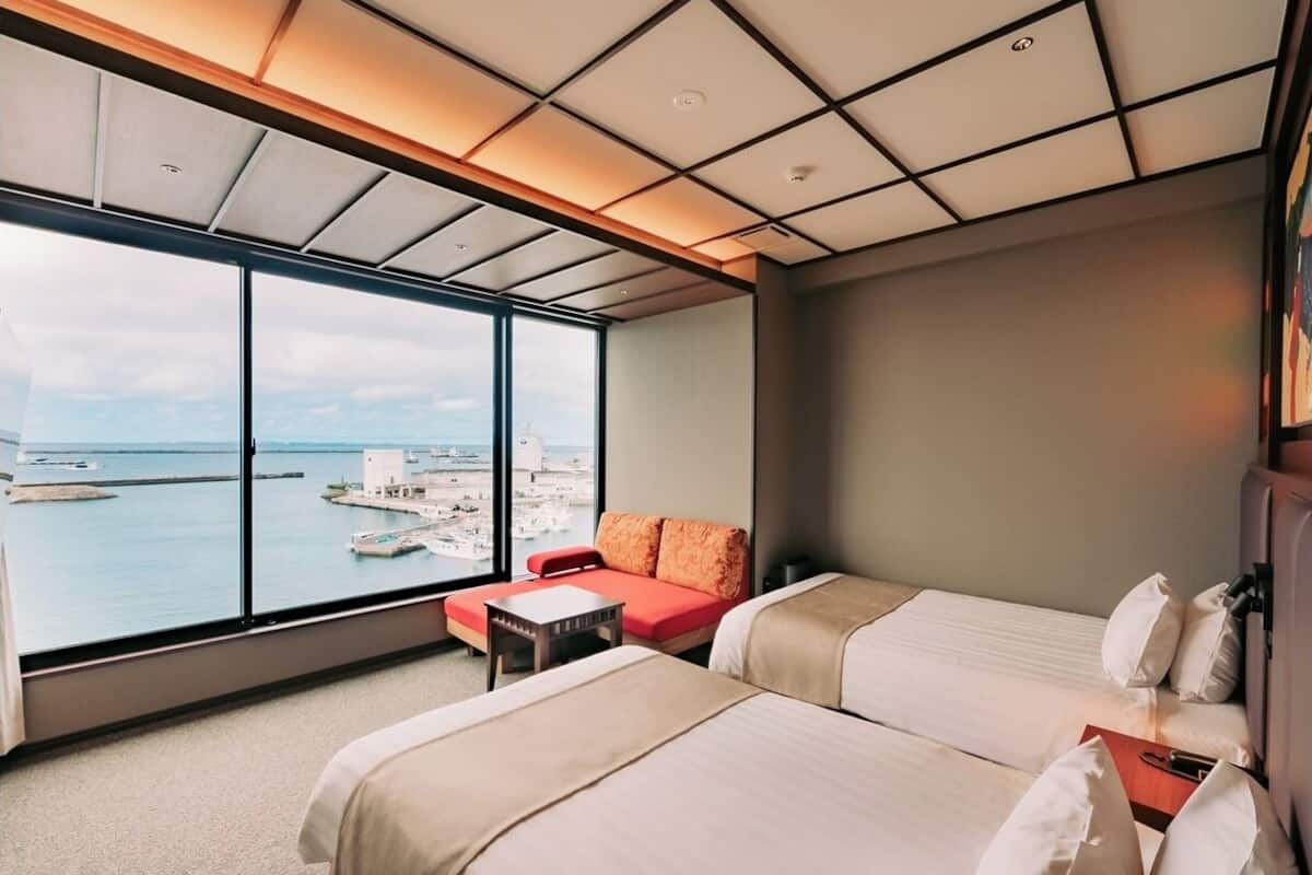 Une chambre d’hôtel avec deux lits, un canapé, une petite table et une grande fenêtre donnant sur la mer et les bateaux.