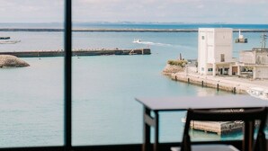 Vista costera desde una habitación con una mesa y una silla, un edificio blanco y un muelle que se adentra en el agua.
