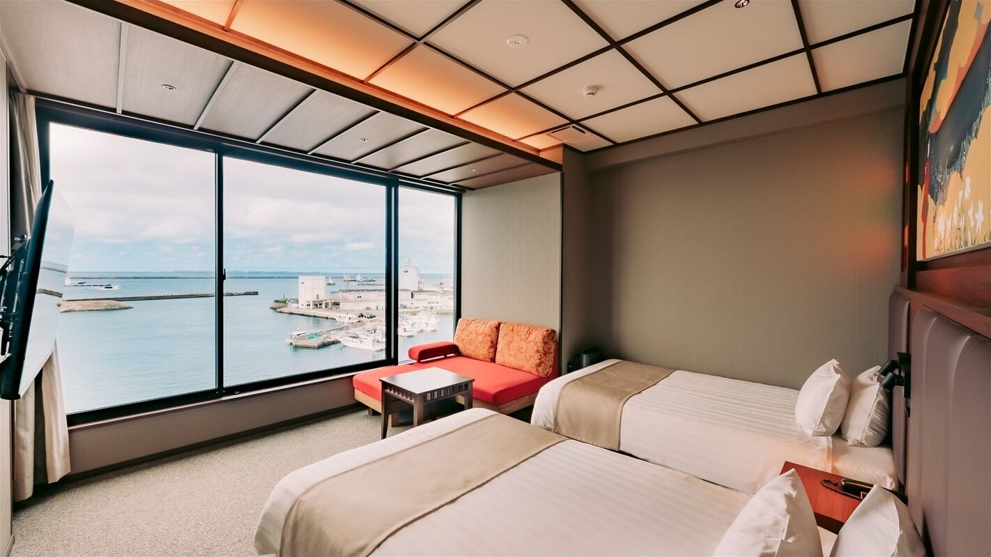 Une chambre d’hôtel avec deux lits, un canapé rouge, une grande fenêtre donnant sur la mer et les bateaux, et un plafond doté d’un éclairage encastré.