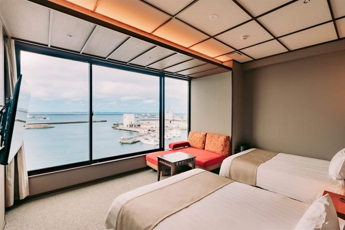 Une chambre d’hôtel avec deux lits, un canapé rouge, une grande fenêtre donnant sur la mer et les bateaux, et un plafond doté d’un éclairage encastré.