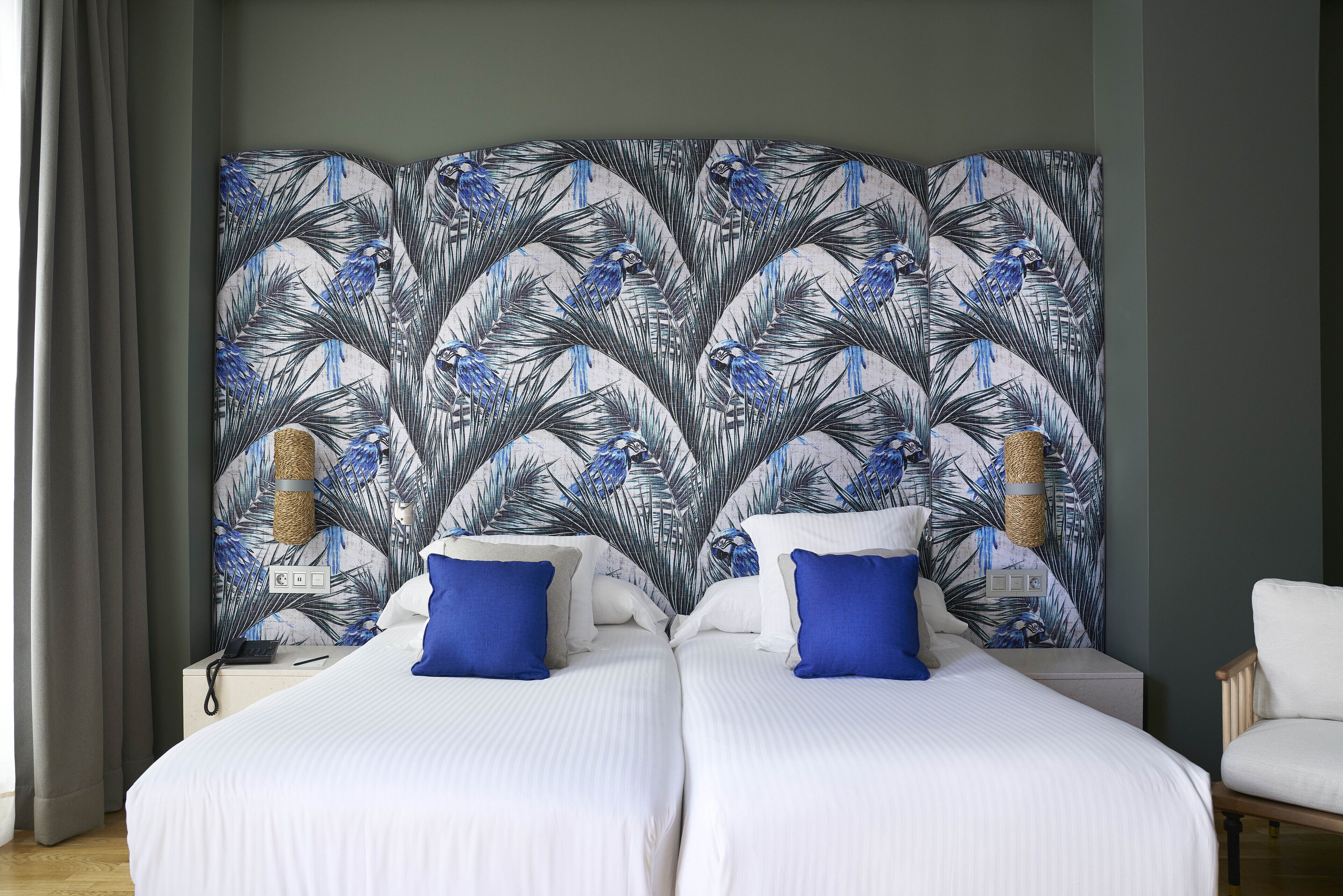 Una habitación de hotel con dos camas, un cabecero de estilo tropical, una mesita de noche con lámpara y una silla.