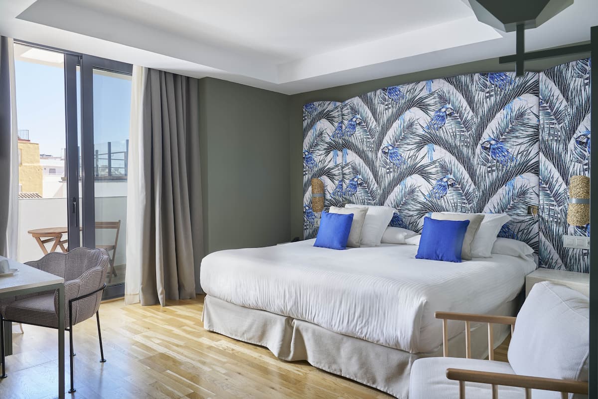 A hotel room with a large bed, two bedside tables, a chair, a balcony with a view, and a tropical leaf-patterned wall.