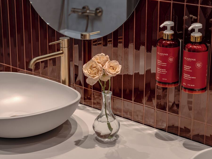 Une salle de bain avec un lavabo blanc, un miroir rond et un vase contenant des fleurs blanches. Des articles de toilette rouges sont posés sur le côté droit.
