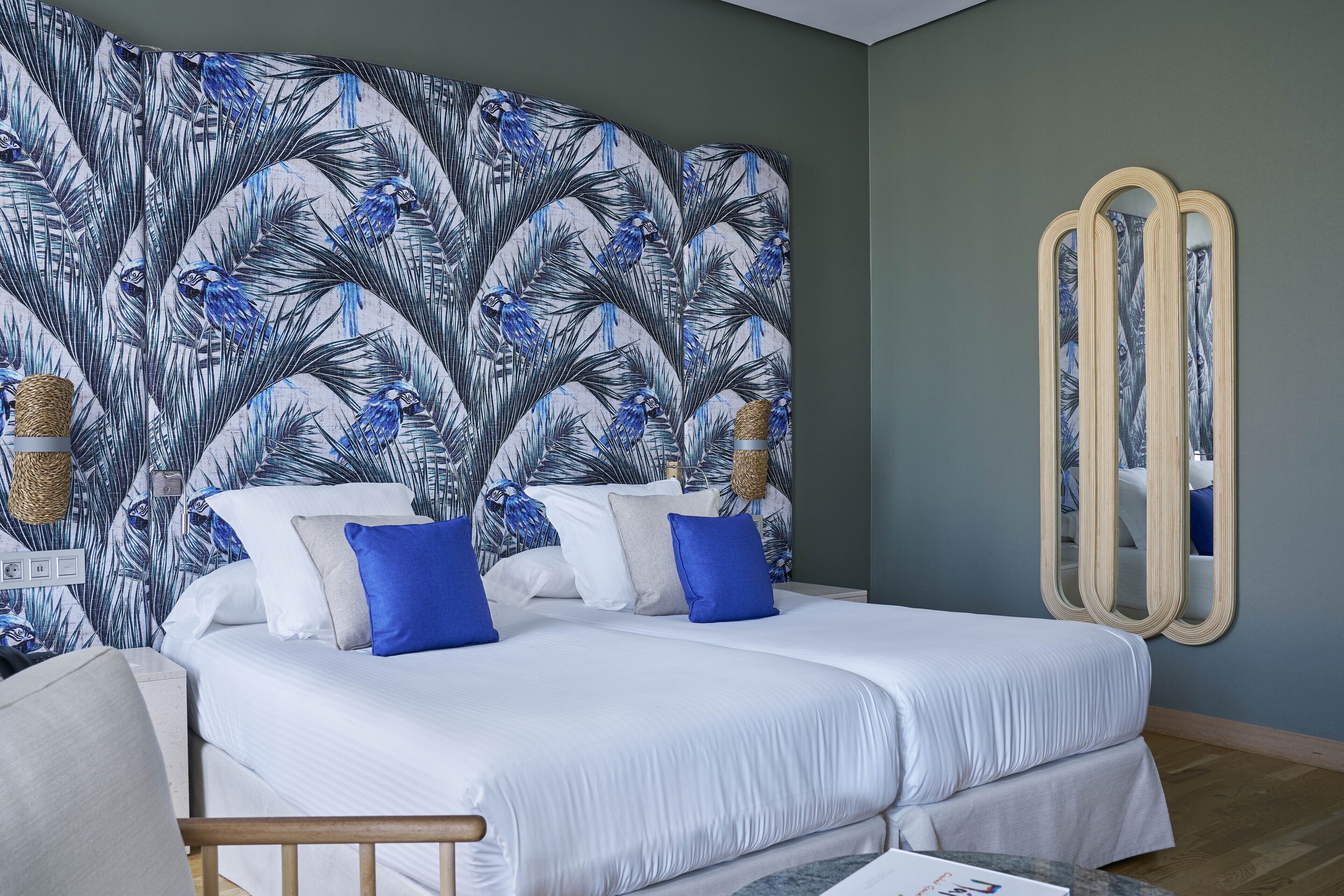 Una cama doble con ropa de cama blanca y almohadas azules, una cesta tejida en la mesita de noche y un espejo en la pared.