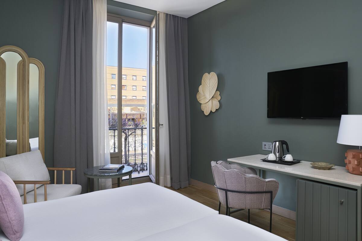 A hotel room with a bed, a desk with a coffee maker, a TV, and a view of a building through the window.