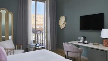 A hotel room with a bed, a desk with a coffee maker, a TV, and a view of a building through a window.