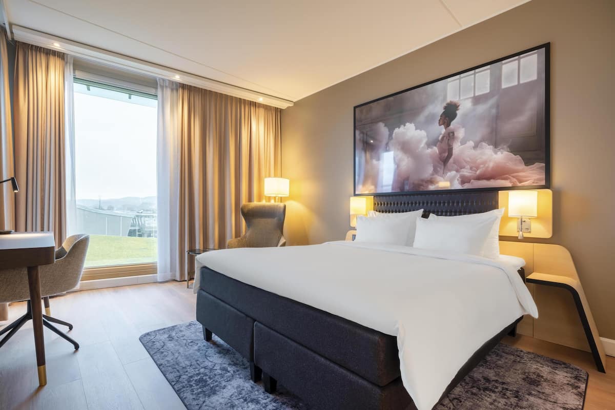 A hotel room with a large bed, a desk, a chair, and a view of the outdoors.