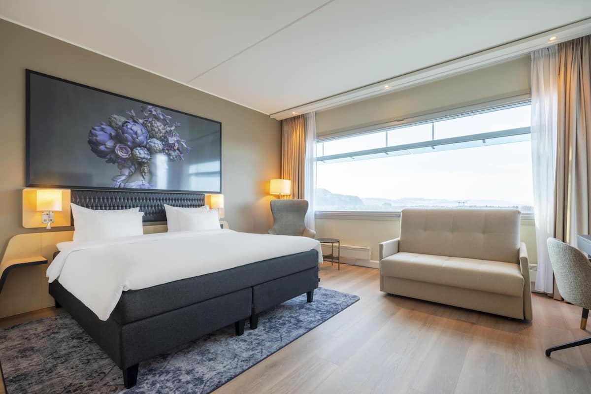 A hotel room with a bed, two chairs, a large window with a view, and a painting on the wall.