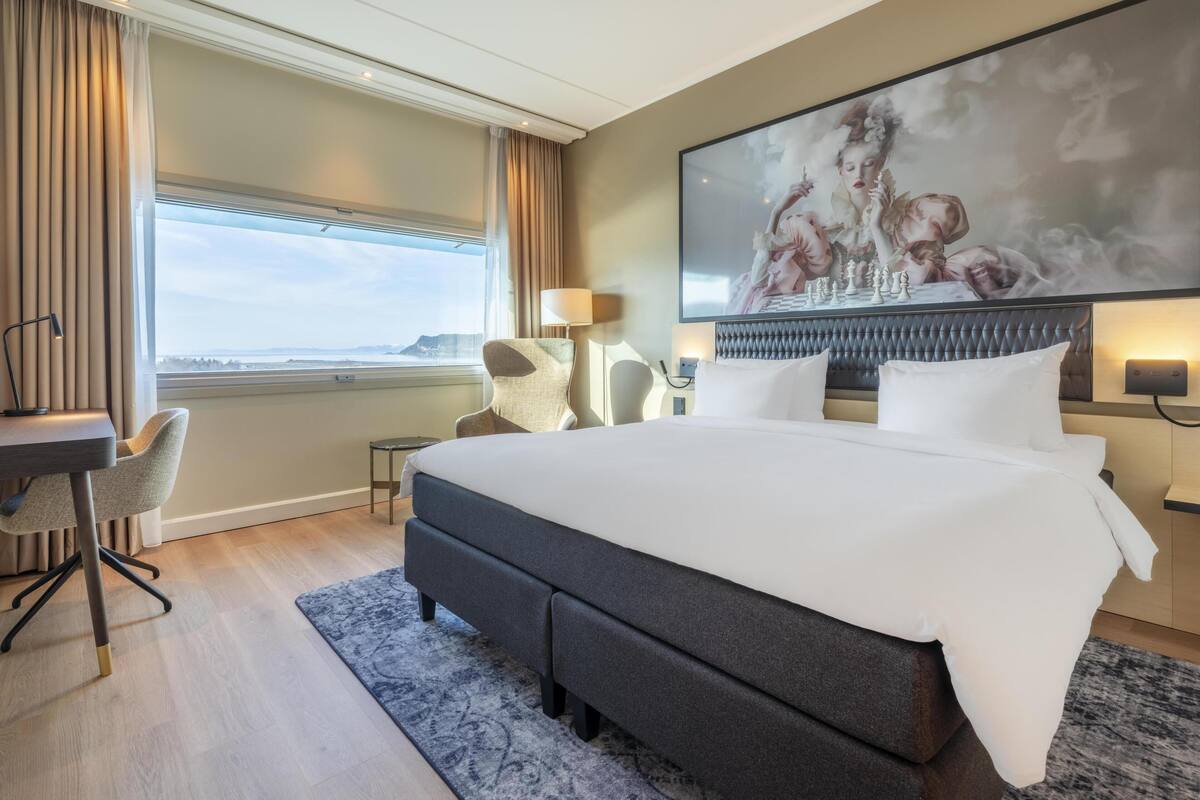 A modern hotel room with a large bed, a desk, a chair, and a large window with a view.