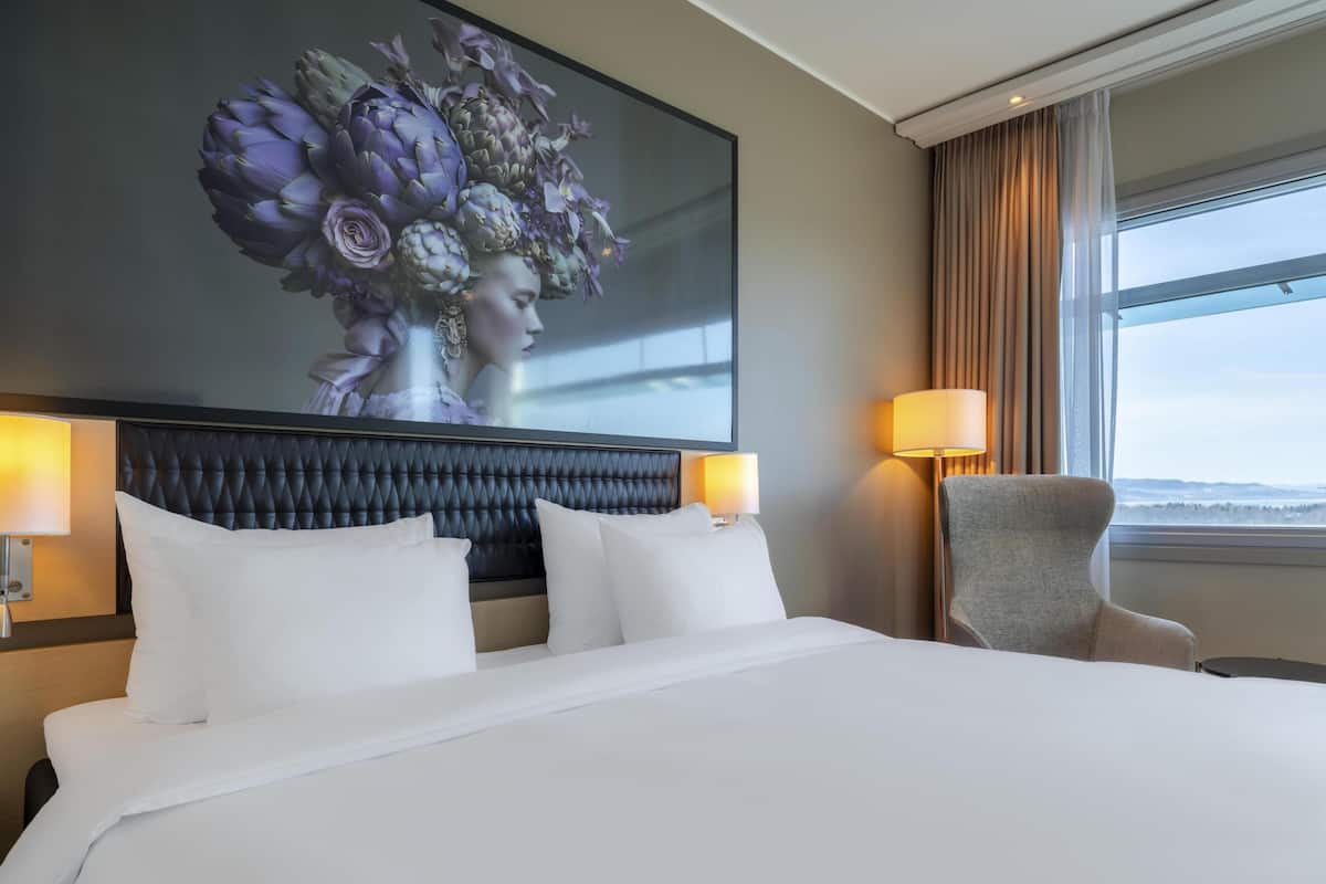 A hotel room with a large bed, two bedside lamps, a chair, and a large window with a view.