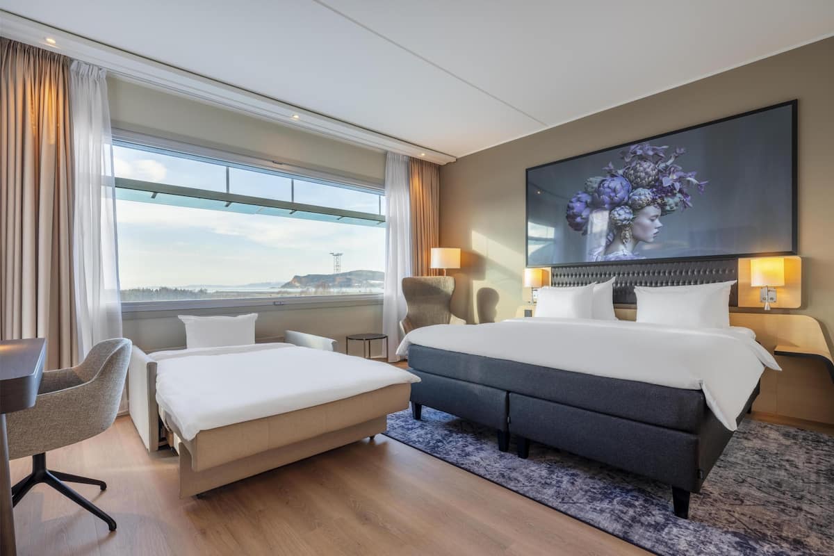 A hotel room with a large bed, a smaller bed, a desk, a chair, a large window with curtains, a painting on the wall, and a view of a distant landscape.
