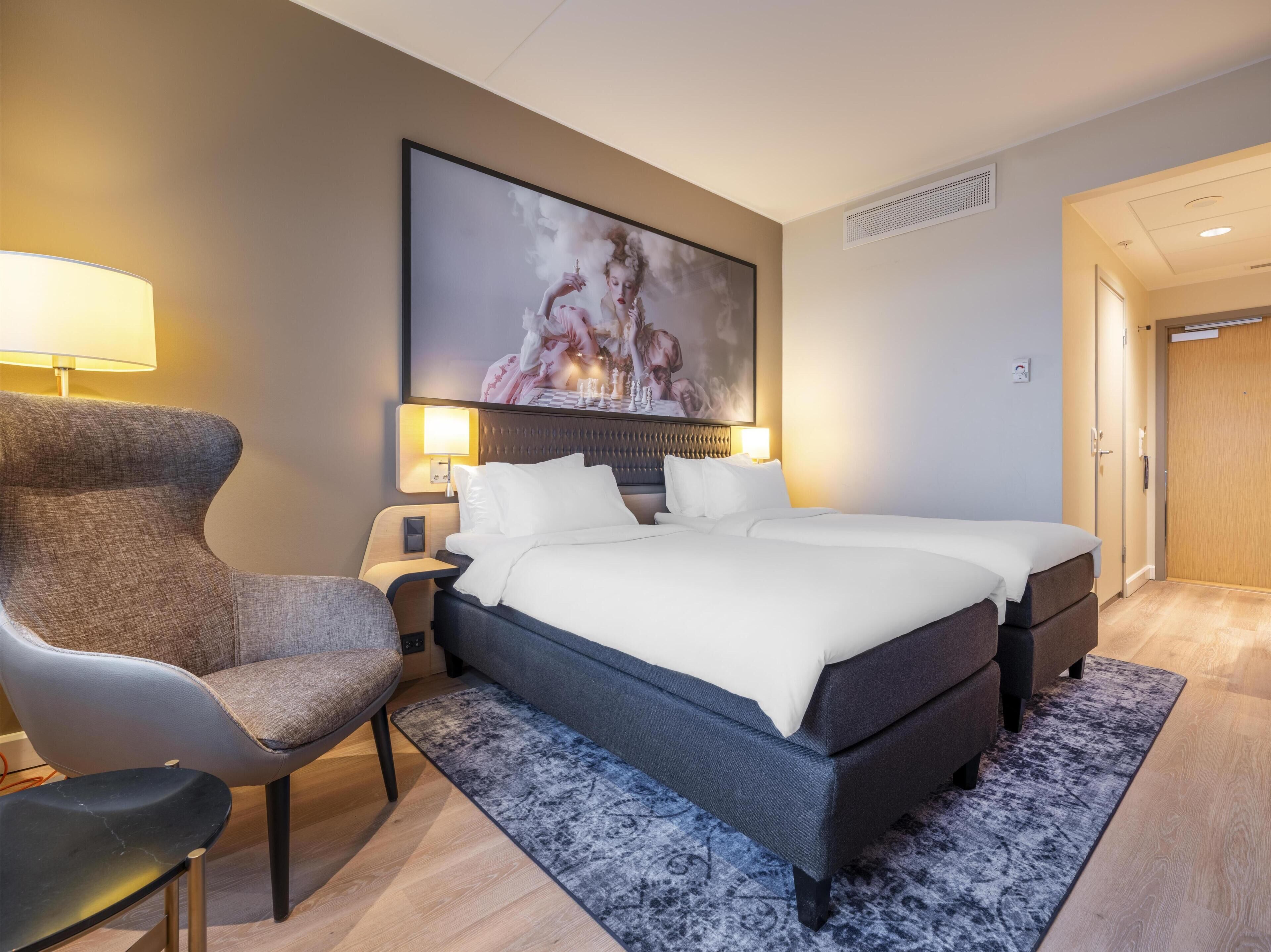 Habitación de hotel con cama, dos mesitas de noche, una silla, un cuadro grande en la pared y una puerta.