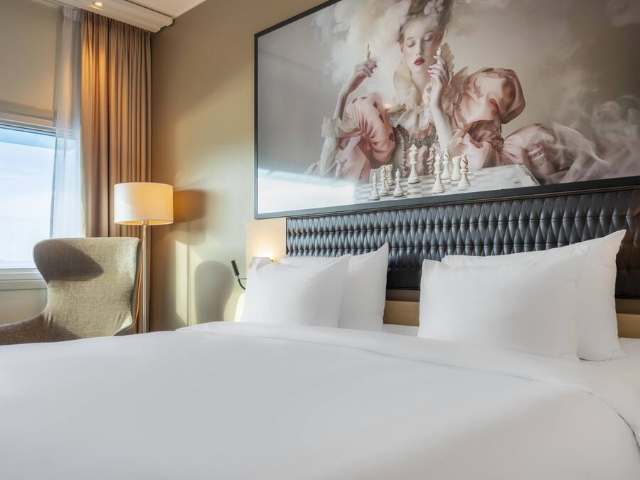 Een hotelkamer met een groot bed, een hoofdbord met een gestuikt ontwerp, een stoel, een lamp en een groot kunstwerk in een lijst aan de muur.
