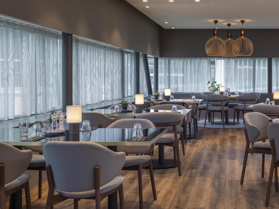 A modern dining area with multiple tables and chairs, large windows with sheer curtains, and pendant lighting.