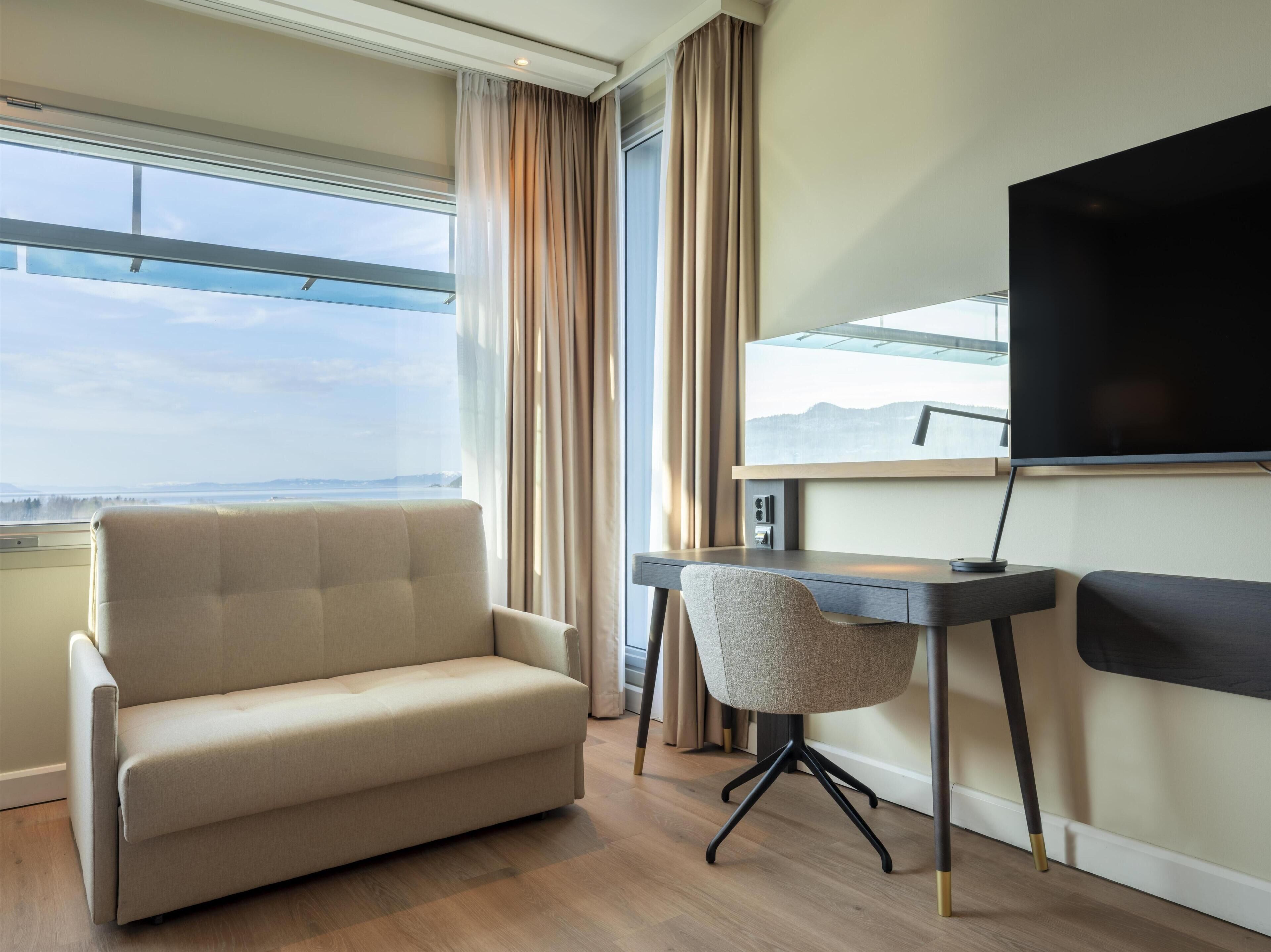 A modern hotel room with a large window, a sofa, a desk, and a flat-screen TV.