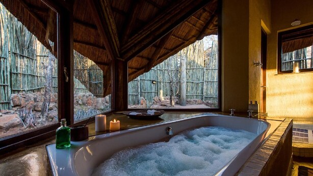 A spa area with a large bathtub filled with bubbles, a bottle of green liquid, and candles on a table. - Tshwene Lodge (Vaalwater)