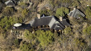 A large, multi-story house with a prominent central chimney, surrounded by dense vegetation and smaller structures. - Tshwene Lodge (Vaalwater)