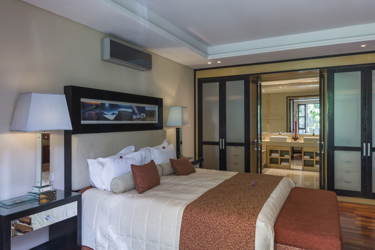 A hotel room with a large bed, two bedside tables with lamps, a framed picture above the bed, and a sliding glass door leading to a bathroom with a vanity and sink.