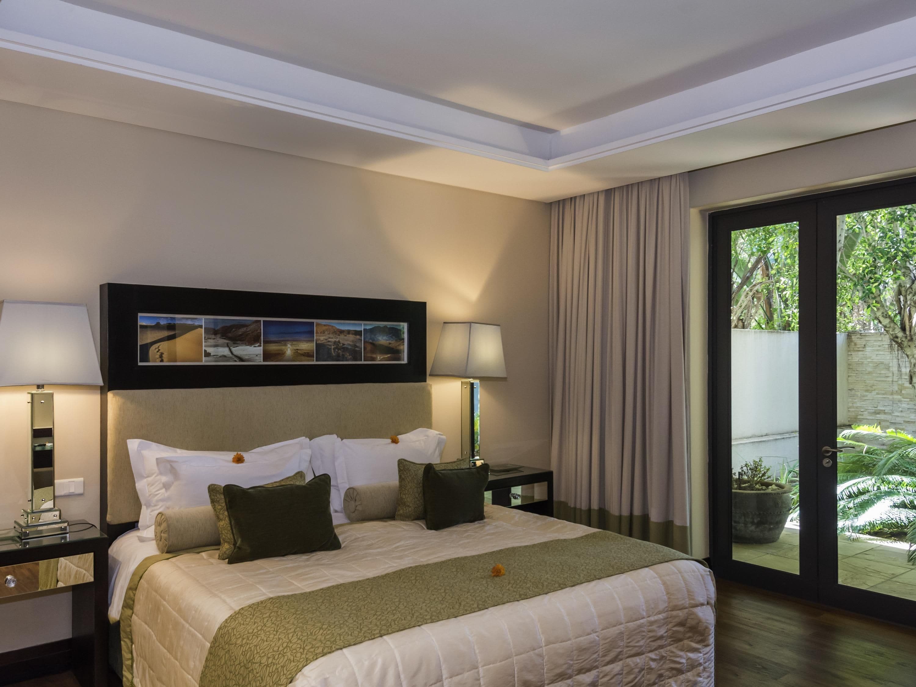 A hotel room with a bed, two bedside tables, a nightstand, a large window with curtains, and a wall-mounted picture.