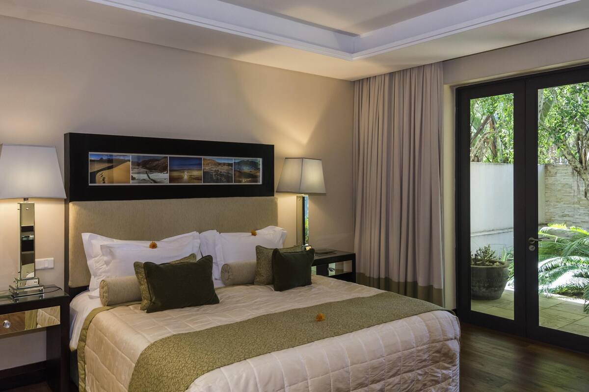 A hotel room with a bed, two bedside tables, a nightstand, a large window with curtains, and a wall-mounted picture.