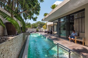 A modern outdoor pool area with a wooden deck, lounge chairs, and a glass-enclosed patio.