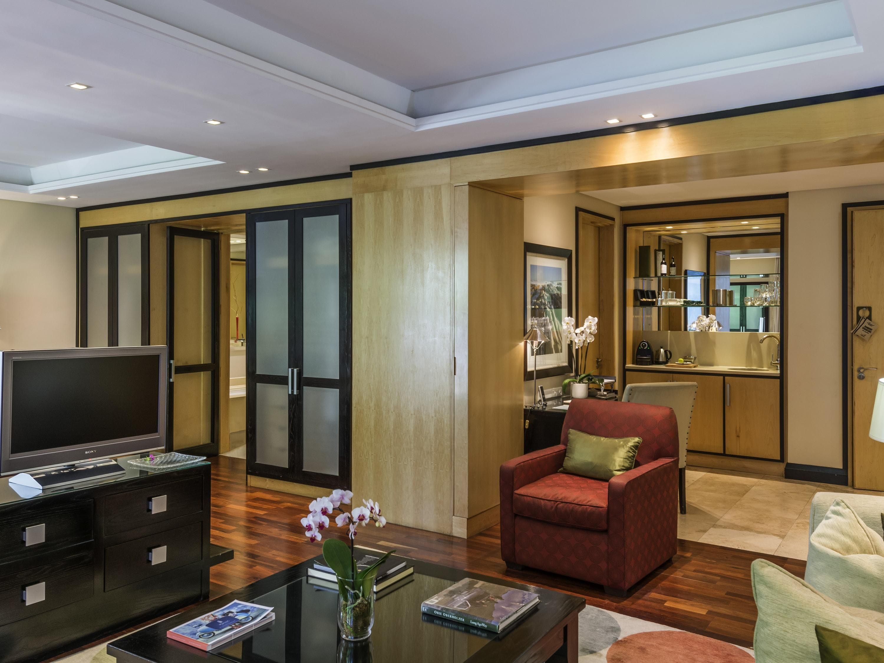 A modern hotel room with a flat-screen TV, a red armchair, a glass coffee table, and a dining area with a kitchen in the background.