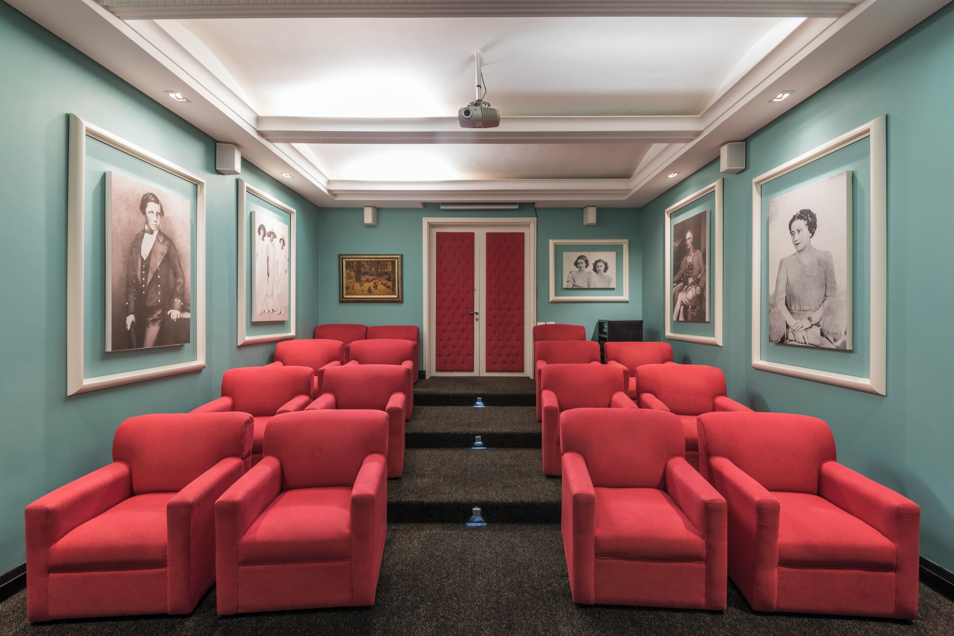 Una sala de teatro con sillas rojas, un proyector y cuadros en las paredes.